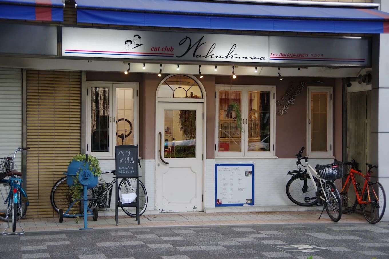 The store has a sign that says "cut club Wakana" on top of a white double door, and offers shortcut and henna services. Sandwiched between the windows and the bike parking lot, there are blue bike racks, houseplants, and menus and signs near the entrance.