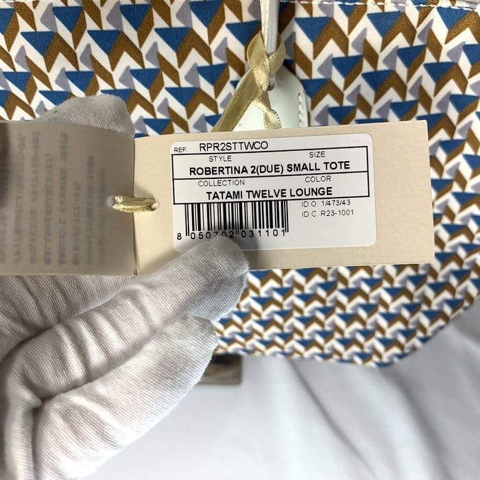 The gloved hand holds the product tag "ROBERTINA 2 (DUE) SMALL TOTE" on top of the tatami pattern fabric with a geometric pattern in blue, brown and white, which is the signature style of ROBERTA PIERI's small tote collection.