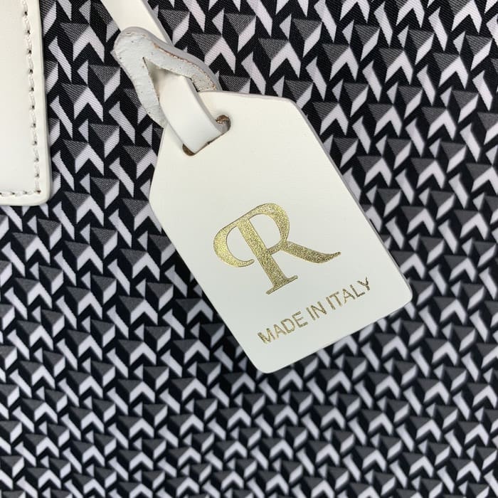 A white leather ROBERTA PIERI tag with the letters "R" in gold and the words "MADE IN ITALY" hangs from the background of a black and white geometric fabric.