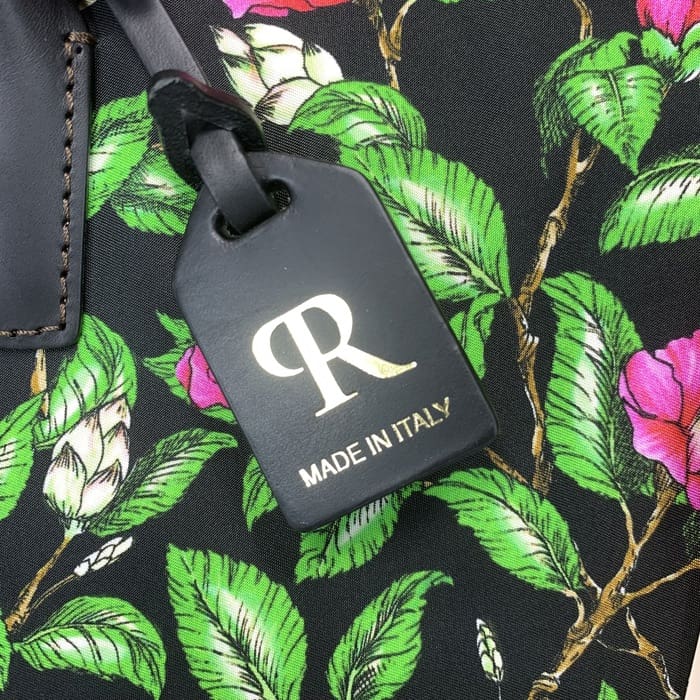 The black ROBERTA PIERI luggage tag with the gold "R" and Italian text is suspended from a floral fabric with green leaves and pink flowers, complementing the mini duffel with timeless Italian elegance.