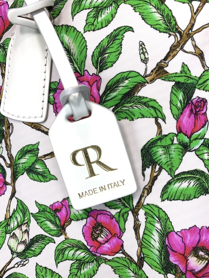 Close-up of ROBERTA PIERI mini duffel bag. peduncles with green leaves and pink flowers; The white tag attached to the 2-way bag features a gold "R" logo and the words "MADE IN ITALY."